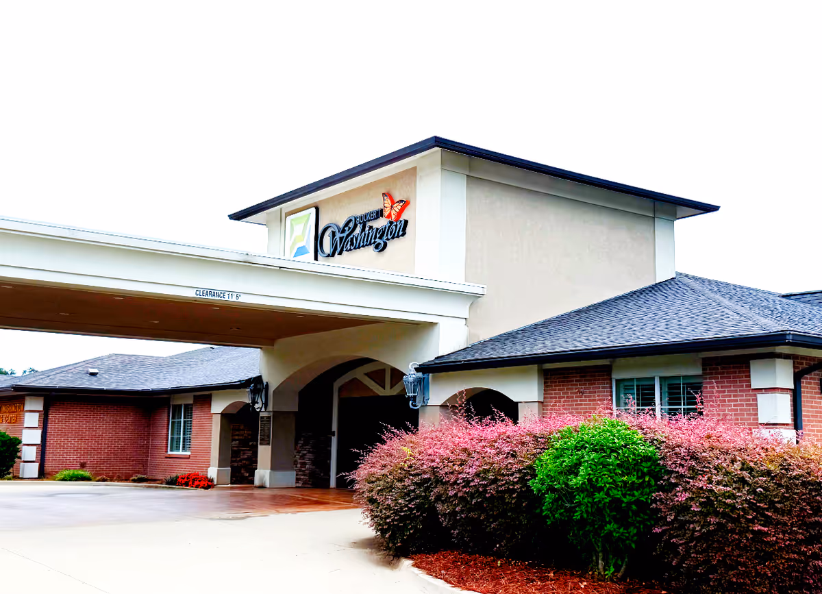 Exterior front view of Booker T. Washington Skilled Nursing and Rehabilitation facility showing the entrance with a covered drop-off area, brick and beige walls, and landscaped bushes with pink and green foliage.