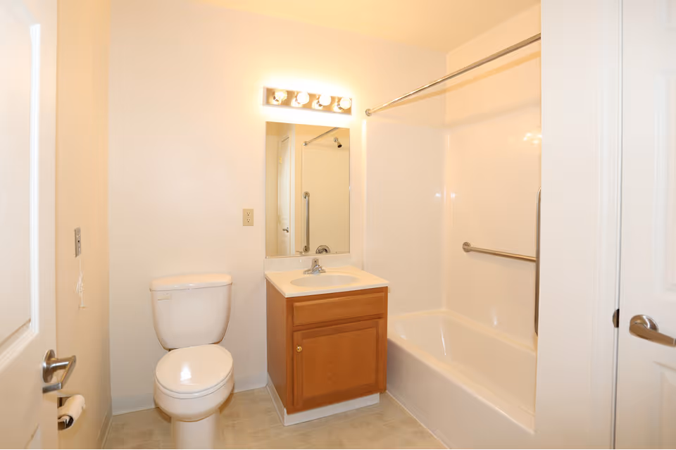 A clean, simple bathroom with a white toilet, a wooden vanity with a sink, a large mirror above the sink with four light bulbs, and a white bathtub with a metal grab bar and shower curtain rod.