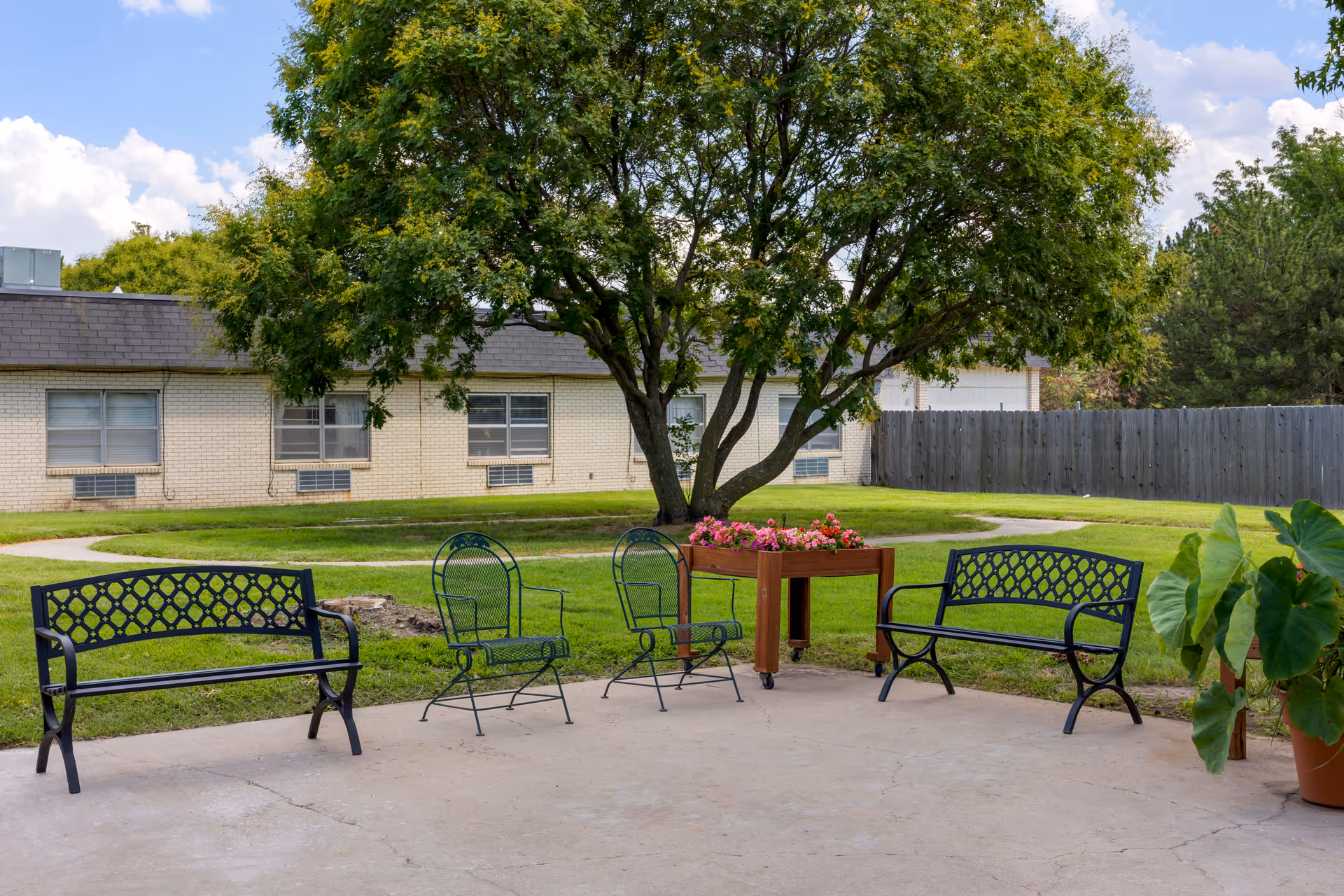 Outdoor patio area with two black metal benches, two green metal chairs, a wooden planter with pink flowers, and a large tree in the background. The area is surrounded by grass, a paved walkway, and a building with windows and a wooden fence.