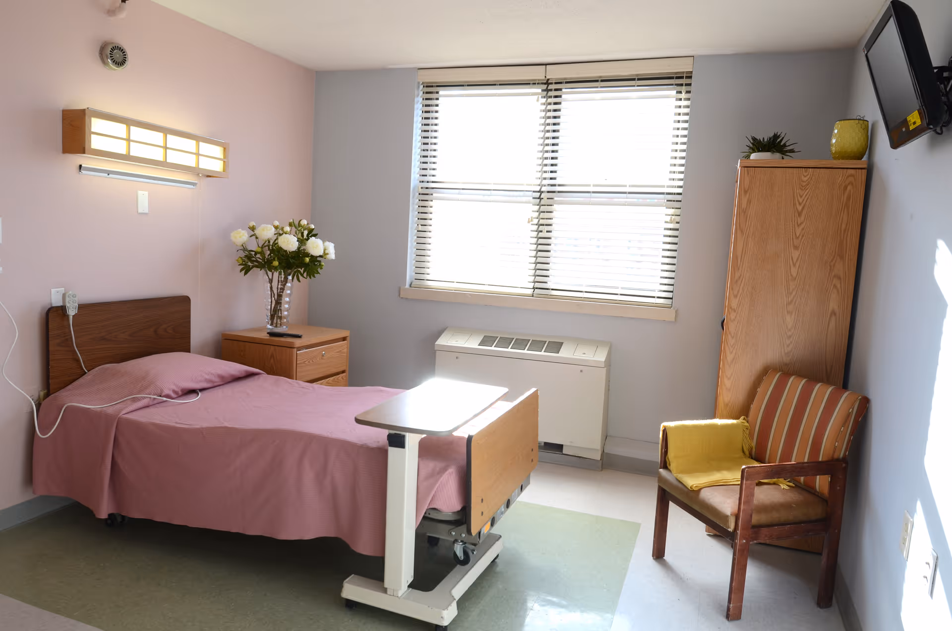 A nursing facility room with a single bed covered in a pink blanket, a wooden nightstand with a vase of white flowers, a window with blinds, a wooden wardrobe, a striped armchair with a yellow blanket, and a wall-mounted television.