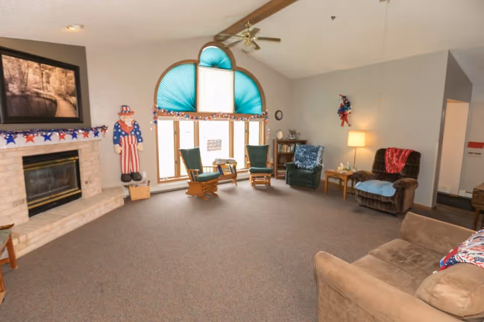 A spacious living room with a beige carpet, a brick fireplace on the left, and a large window with teal fan-shaped blinds. The room is decorated with patriotic red, white, and blue garlands and a wooden Uncle Sam figure. There are several chairs including two green rocking chairs, a green armchair, a brown recliner with a red blanket, and a beige sofa with a patriotic pillow. A small wooden table with a lamp and decorations is next to the recliner.
