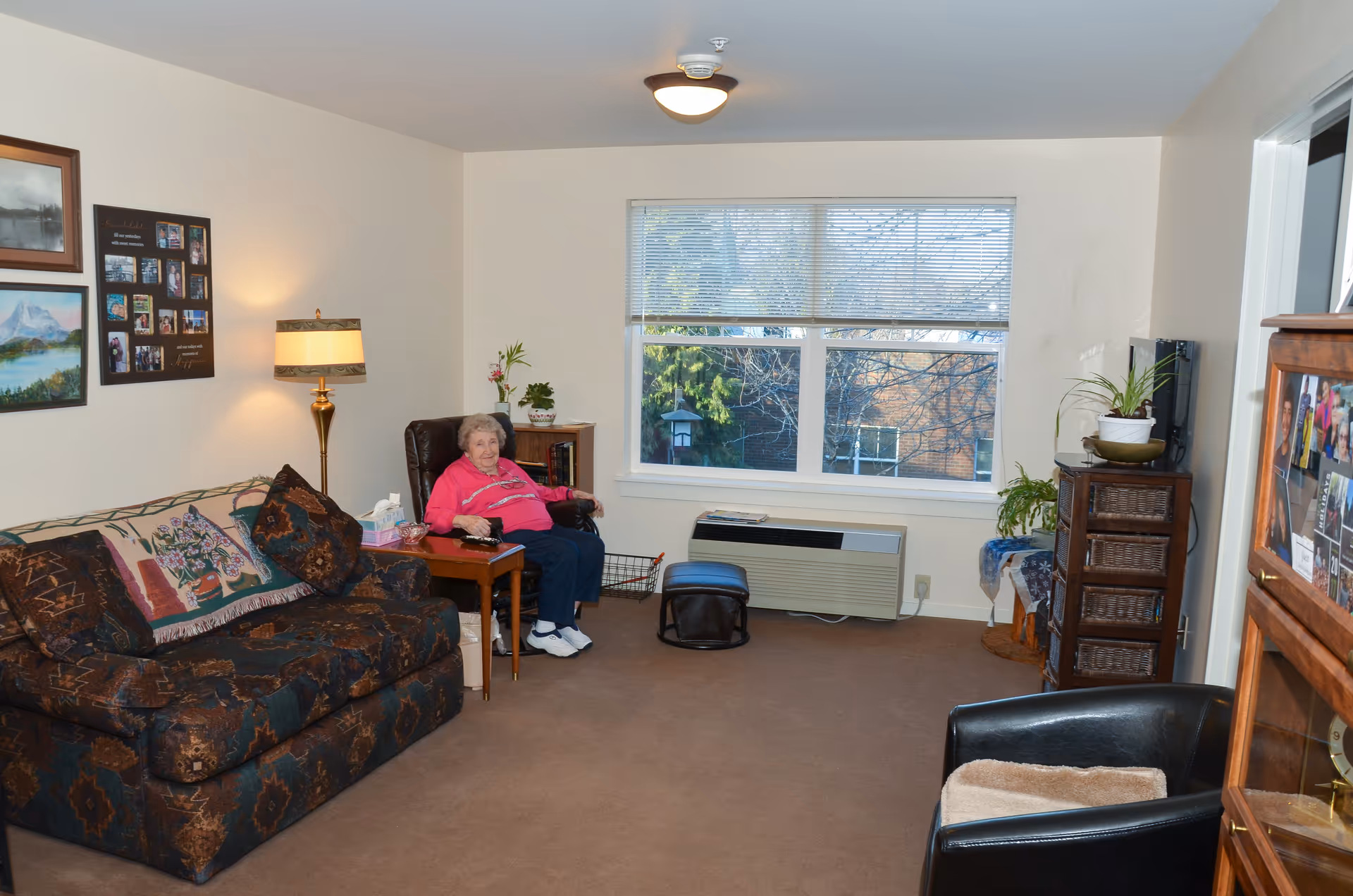 A cozy living room in a senior living facility with a large window showing trees outside. An elderly woman in a pink sweater sits in a brown recliner chair next to a wooden side table with a lamp and tissue box. There is a patterned sofa with pillows and a blanket, a black armchair with a cushion, and various plants and framed pictures on the walls.