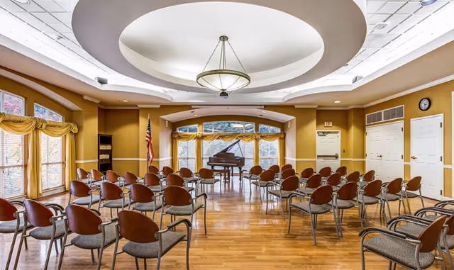 Large multipurpose activity room with rows of chairs facing a grand piano beneath a circular recessed ceiling fixture.