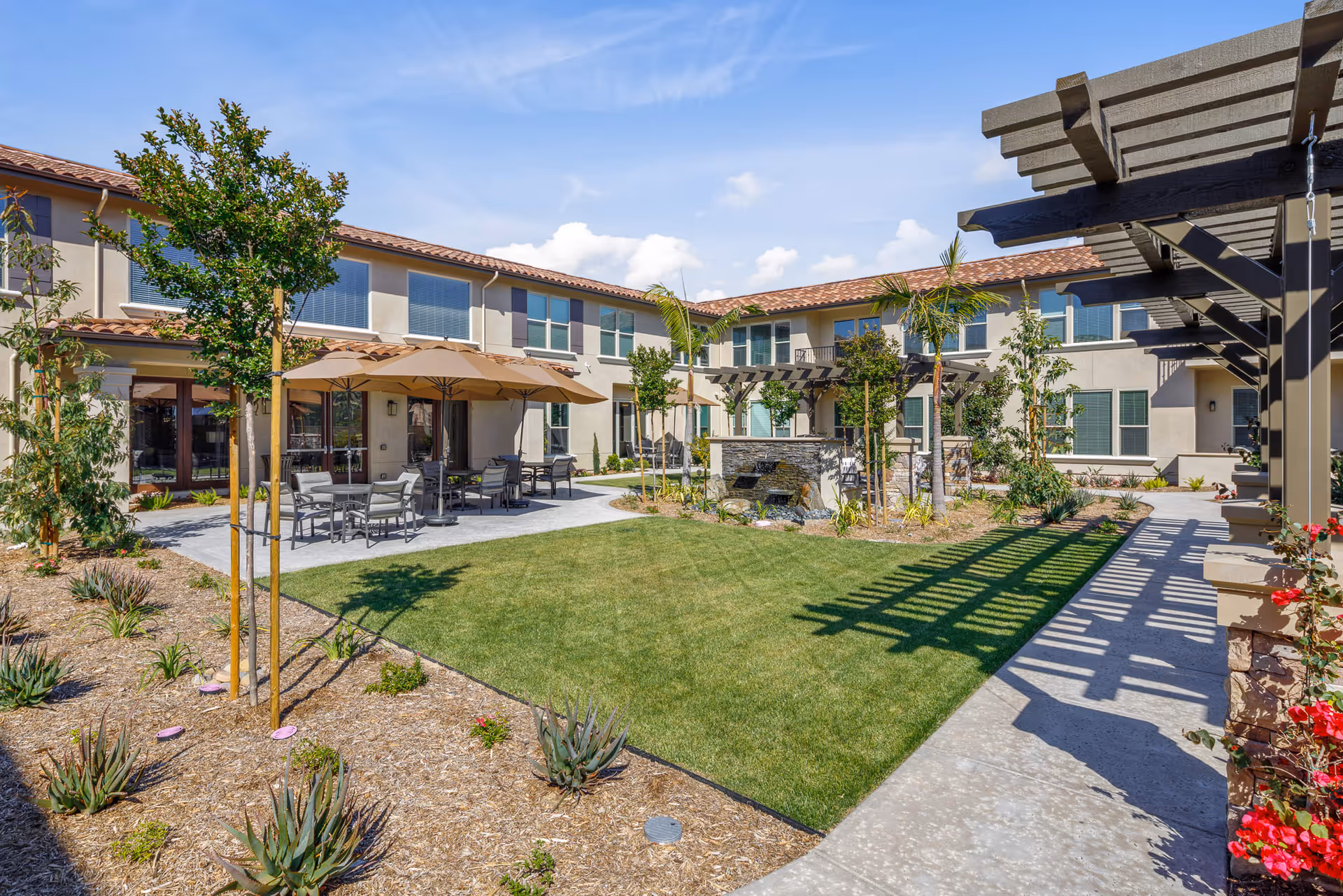 Outdoor courtyard area of a senior living facility with green lawn, small trees, shrubs, patio tables with umbrellas, and a pergola casting shadows on a walkway. The building surrounding the courtyard has two stories with multiple windows and a tiled roof under a blue sky with some clouds.