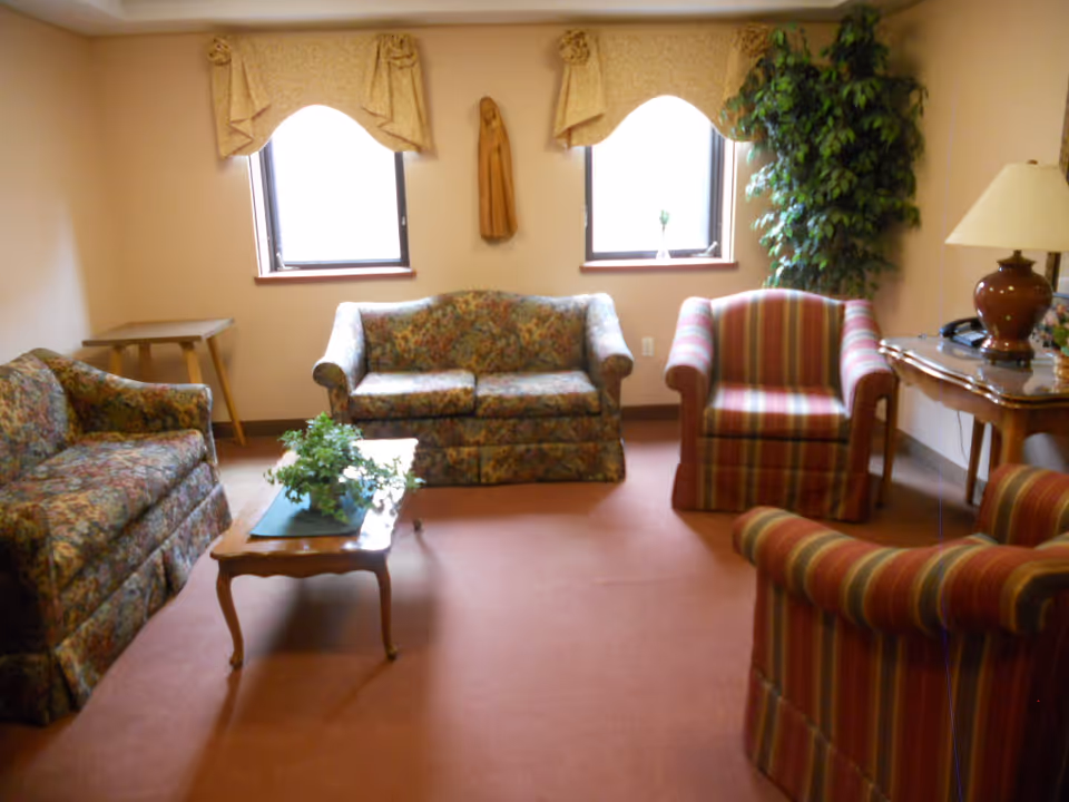 A cozy living room with two floral-patterned sofas, two striped armchairs, a wooden coffee table with a green plant, a side table with a lamp and telephone, two windows with beige valances, and a tall green plant in the corner.