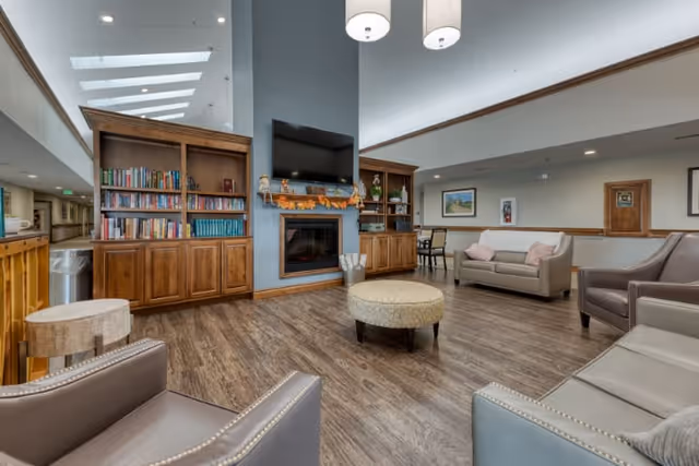 A spacious and well-lit living room area in a senior living facility featuring a central fireplace with a TV mounted above it, flanked by wooden bookshelves filled with books and decorative items. The room has multiple comfortable armchairs and sofas arranged around a round ottoman on a wood floor. The ceiling is high with skylights and modern hanging light fixtures. There is a hallway visible to the left and a dining area with tables and chairs in the background.