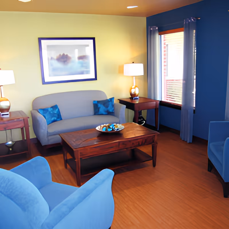 A cozy living room with a gray sofa adorned with blue pillows, two blue armchairs, a wooden coffee table with a decorative bowl, two side tables each with a lamp, and a window with light curtains. The walls are painted yellow and blue.