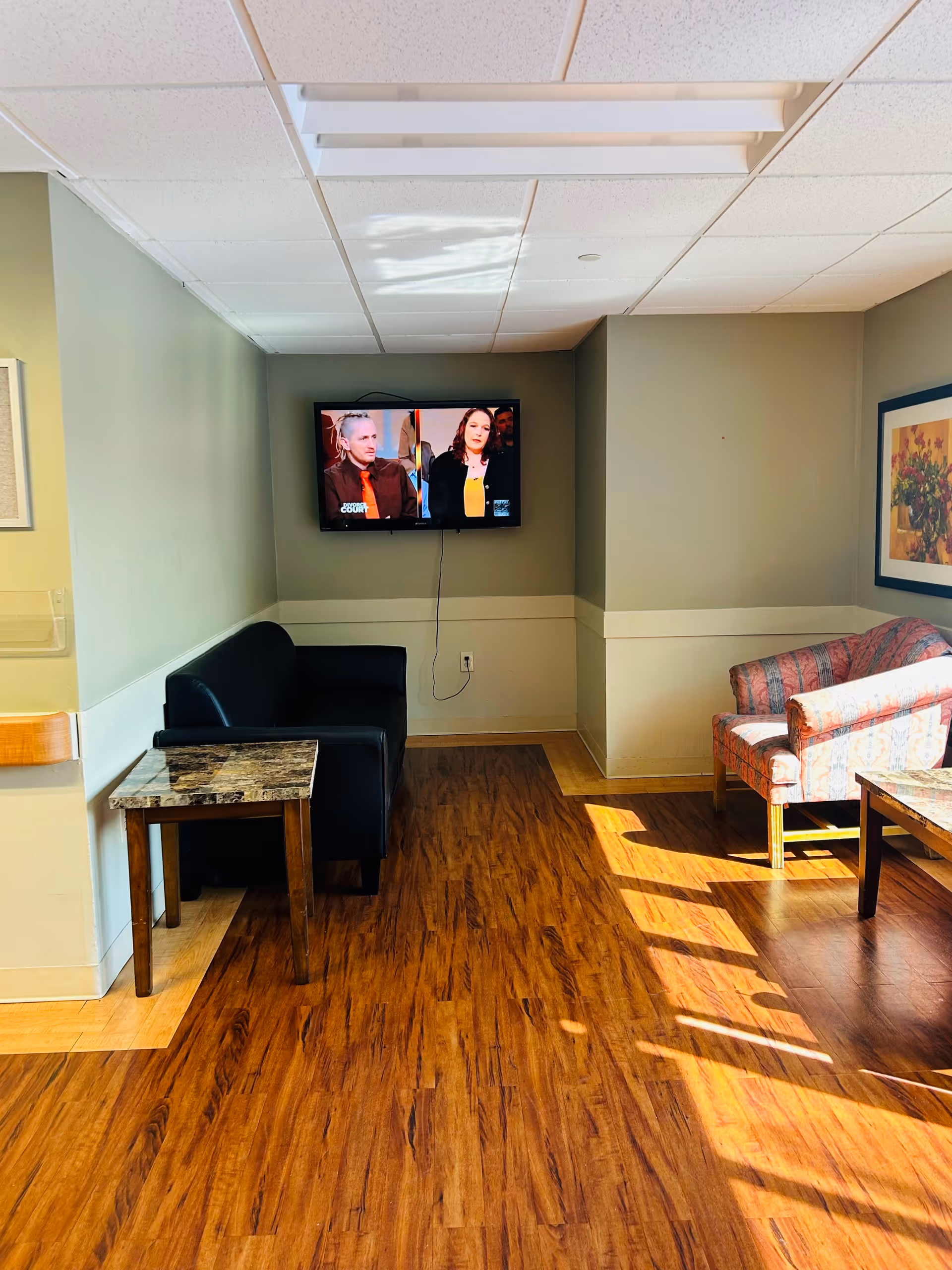 Small common lounge with a wall-mounted TV, a black loveseat, patterned chairs, side tables, framed art, and wood-look flooring.