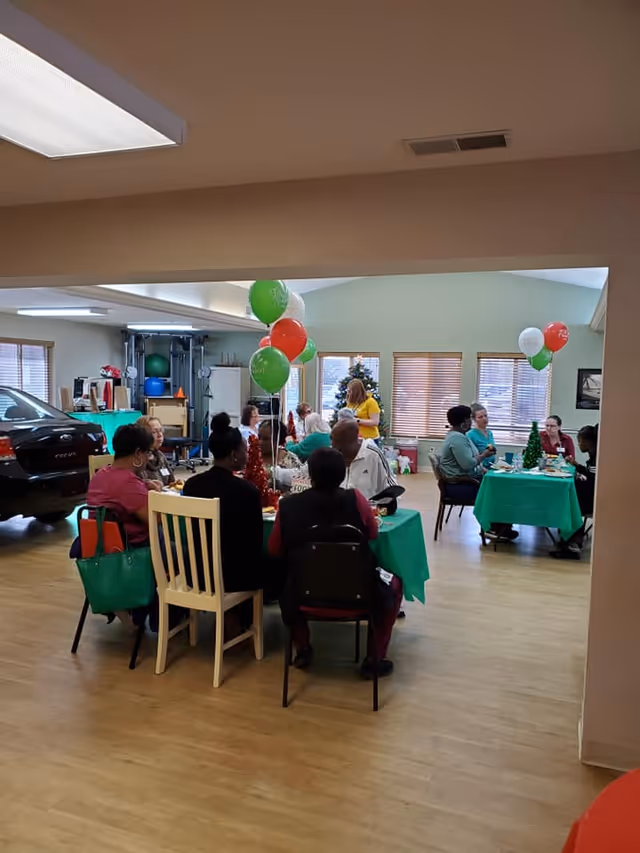 A group of people sitting around tables covered with green tablecloths in a spacious room decorated with red, green, and white balloons. There is a Christmas tree in the background and some holiday decorations on the tables. The room has wooden flooring, windows with blinds, and exercise equipment in one corner.