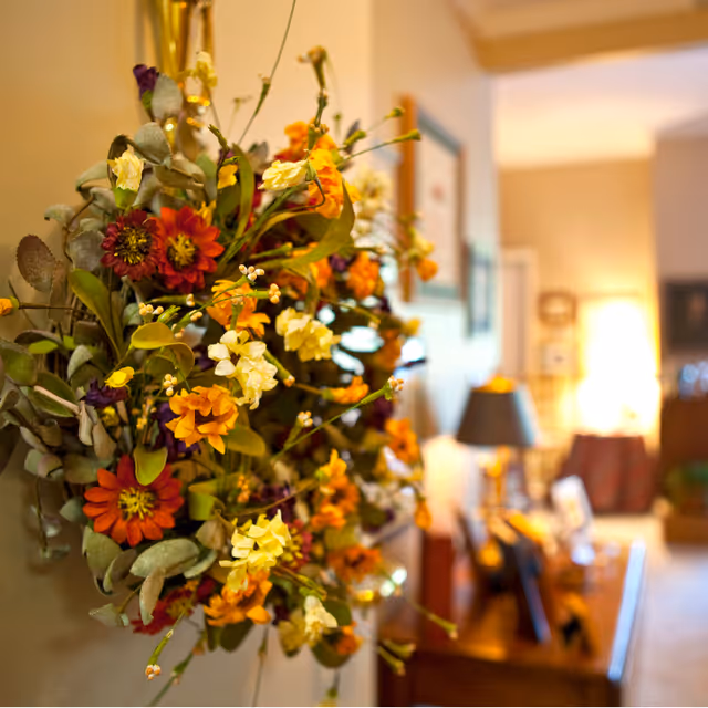 Close-up of a colorful floral wreath with orange, yellow, and purple flowers hanging on a wall inside a warmly lit room. In the background, there is a wooden table with a lamp and framed pictures, and the room appears cozy and inviting.