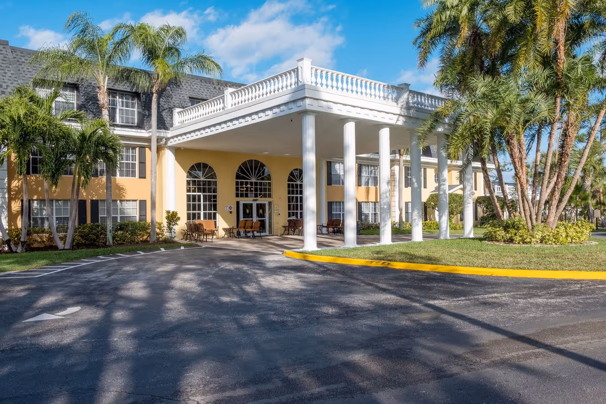 Front exterior view of Volante Senior Living of Delray Beach building with a covered entrance supported by white columns, surrounded by palm trees and landscaping under a blue sky.