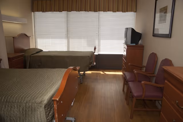 A senior living facility bedroom with two single beds covered in green bedspreads, a wooden nightstand between the beds, two purple cushioned chairs with wooden armrests, a wooden dresser with a television on top, and a large window with closed blinds and a valance.