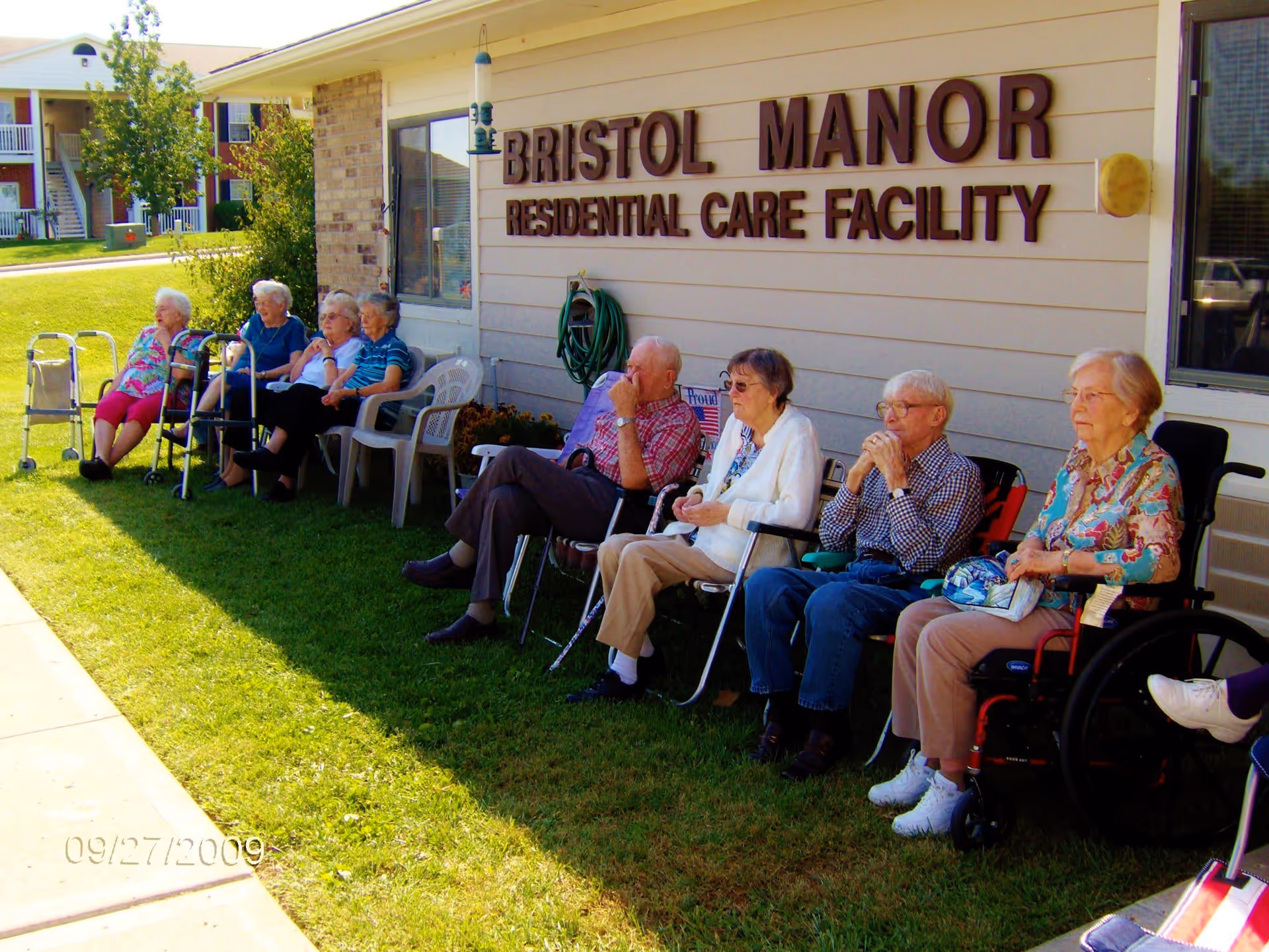 A group of elderly people sitting outside on chairs and wheelchairs along the side of a building with a sign that reads 'Bristol Manor Residential Care Facility'. The scene is sunny with green grass and a sidewalk nearby.
