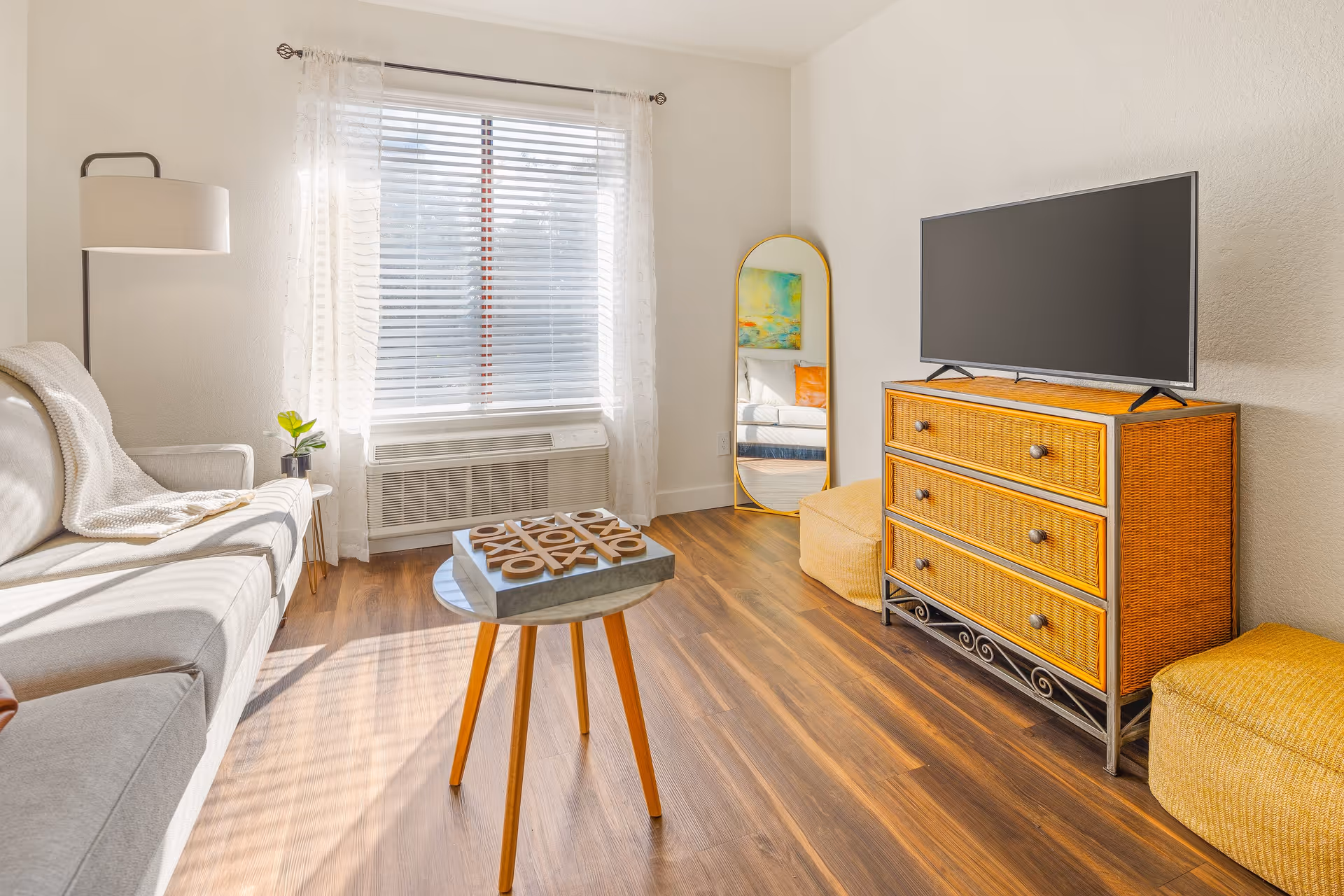 A bright living room with a large window covered by sheer white curtains and blinds. The room features a light gray sofa with a white throw blanket, a small round wooden coffee table with a tic-tac-toe game on it, a floor lamp, a wicker dresser with a flat-screen TV on top, a tall oval mirror with a gold frame, and two yellow poufs on a wood floor.