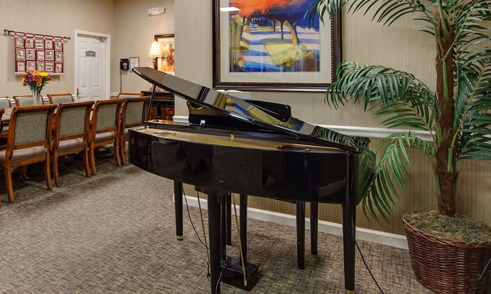 Interior room with a black grand piano in the foreground, a large potted plant to the right, a long wooden table with chairs along the left wall, and a colorful framed painting on the wall behind the piano.