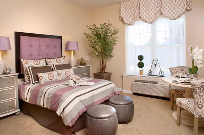 Well-decorated bedroom with a purple-upholstered headboard, striped bedding, two bedside tables, two round ottomans, and a window-side table and chairs.
