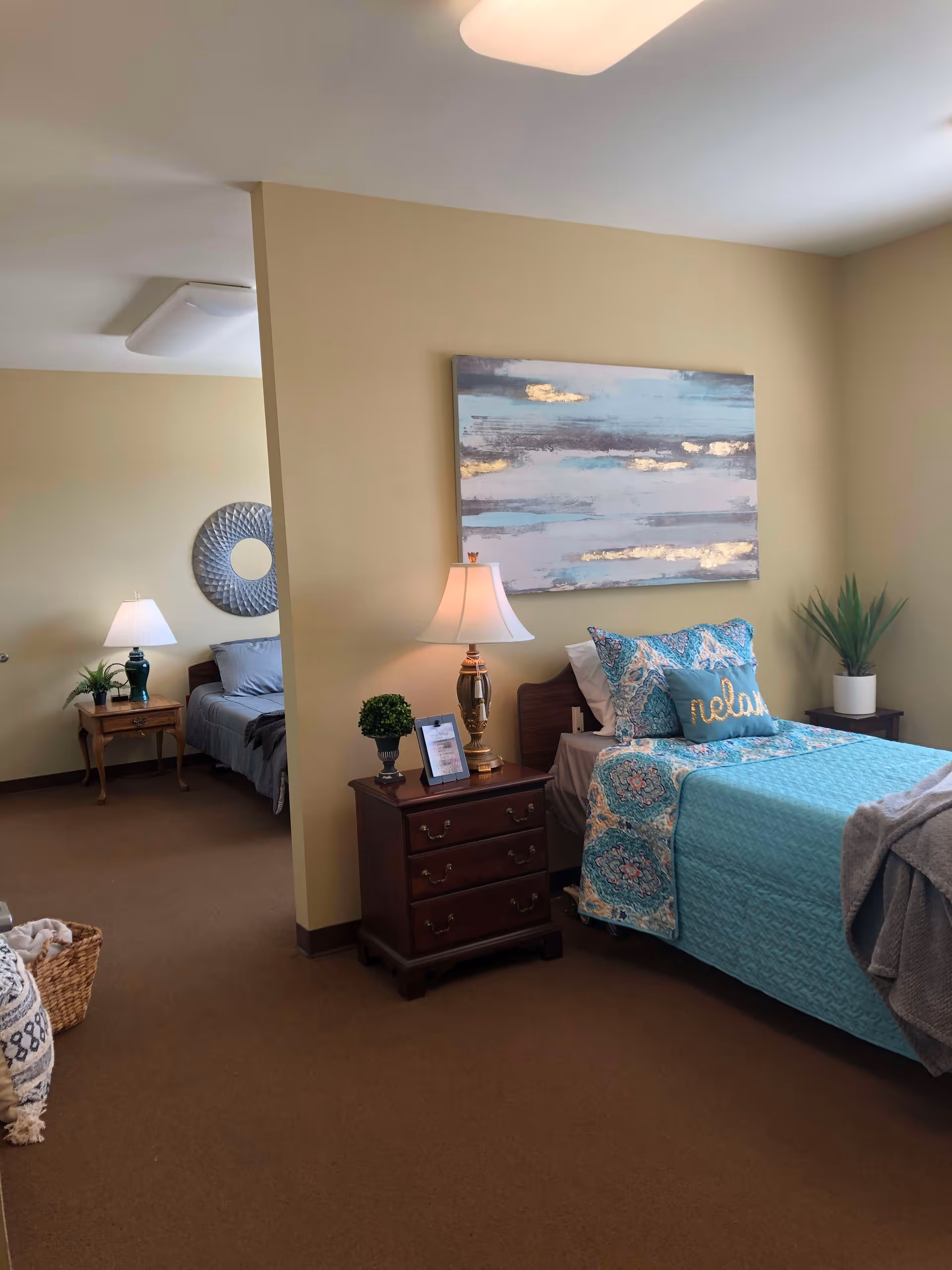 A cozy bedroom in a memory care facility featuring a single bed with blue and patterned bedding, a pillow with the word 'relax', a wooden nightstand with a lamp, a small plant, and a framed picture. In the background, there is another bed with gray bedding, a wooden side table with a lamp and plant, and a decorative round mirror on the wall. The walls are painted beige and the carpet is brown.