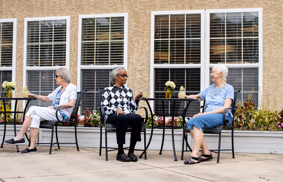 Three elderly women sitting outside at two black metal tables with drinks in their hands, positioned in front of a beige building with large windows and flower boxes beneath them.