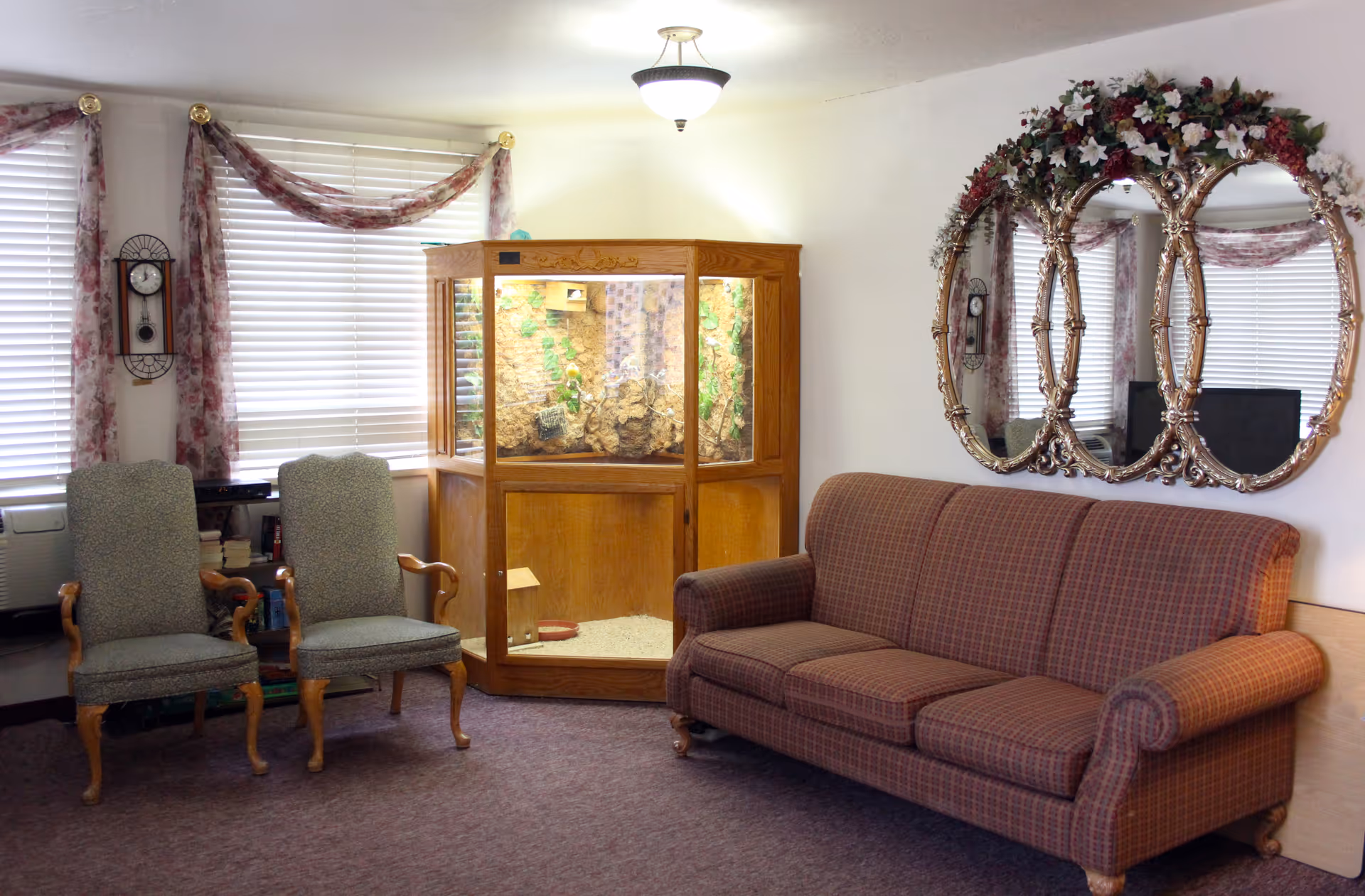 A cozy living room area with a patterned three-seat sofa on the right and two upholstered armchairs with wooden arms on the left. Between the chairs and sofa is a large wooden terrarium or display case with plants inside. The walls are white, decorated with a large ornate triple oval mirror adorned with floral decorations above the sofa. The windows have white blinds and floral curtains draped with a swag style. A wall clock is mounted between the windows.