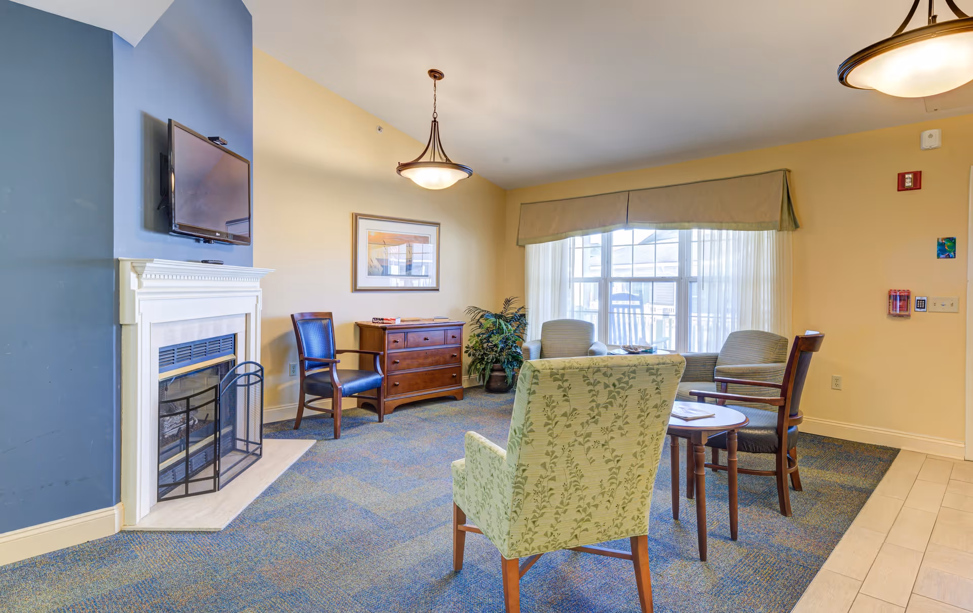A cozy senior living community lounge area with a fireplace and a wall-mounted TV above it. The room features a mix of chairs including a patterned armchair, wooden chairs with cushions, and a small round table. There is a large window with sheer curtains and a valance letting in natural light. A wooden dresser and a potted plant are placed against the wall, and a framed picture hangs above the dresser. The walls are painted in soft yellow and blue tones.