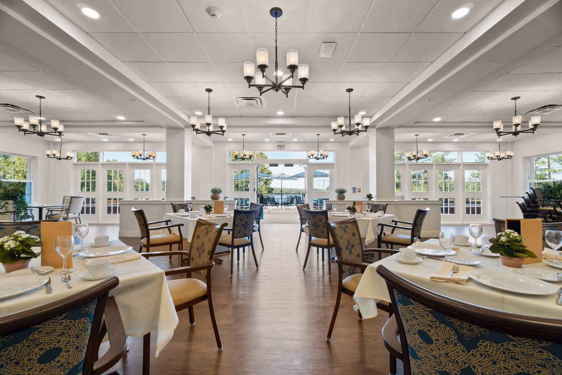 A bright and spacious dining room in a senior living facility with multiple tables set with white tablecloths, plates, cups, and glassware. The room features wooden chairs with patterned upholstery, large windows and glass doors letting in natural light, and several chandeliers hanging from the ceiling. Outside, there is a patio area with umbrellas visible through the glass doors.