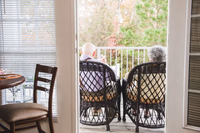 Two elderly people sitting on wicker chairs on a balcony or porch, viewed from inside a room. The balcony overlooks greenery and trees, and there is a table and chair visible inside the room near a window with blinds.