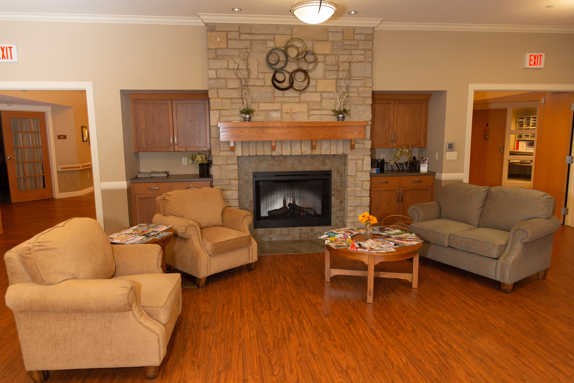 A cozy living room area in a senior living facility with a stone fireplace in the center. There are two beige armchairs on the left and a gray sofa on the right, arranged around a wooden coffee table with magazines and a small flower vase. Wooden cabinets flank the fireplace, and there are two exit signs visible above doorways on either side of the room. The floor is a warm-toned wood laminate.