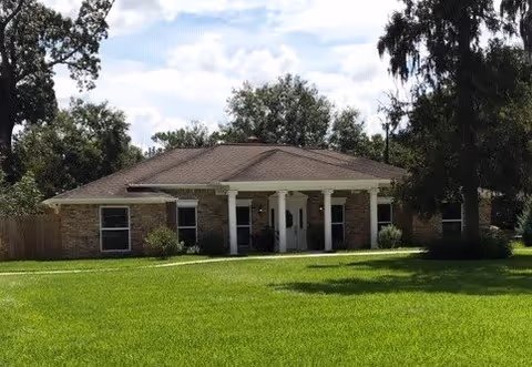 Single-story brick building with a brown roof and white columns at the entrance, surrounded by green grass and trees under a partly cloudy sky.