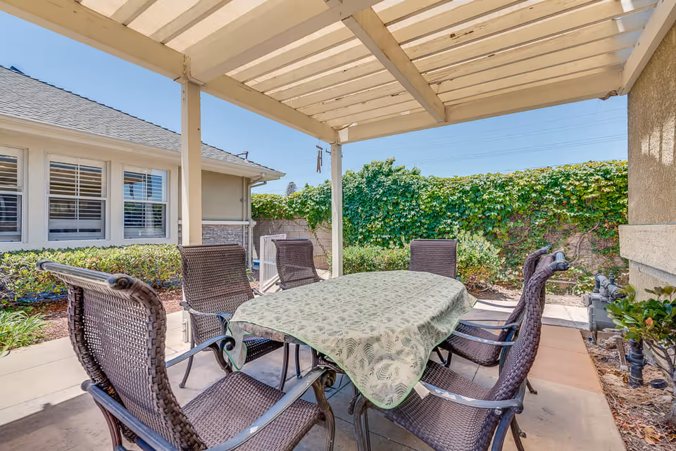 Outdoor patio area with a rectangular table covered by a green patterned tablecloth surrounded by six brown wicker chairs. The patio is shaded by a pergola and is adjacent to a building with windows and surrounded by green bushes and a tall leafy hedge under a clear blue sky.