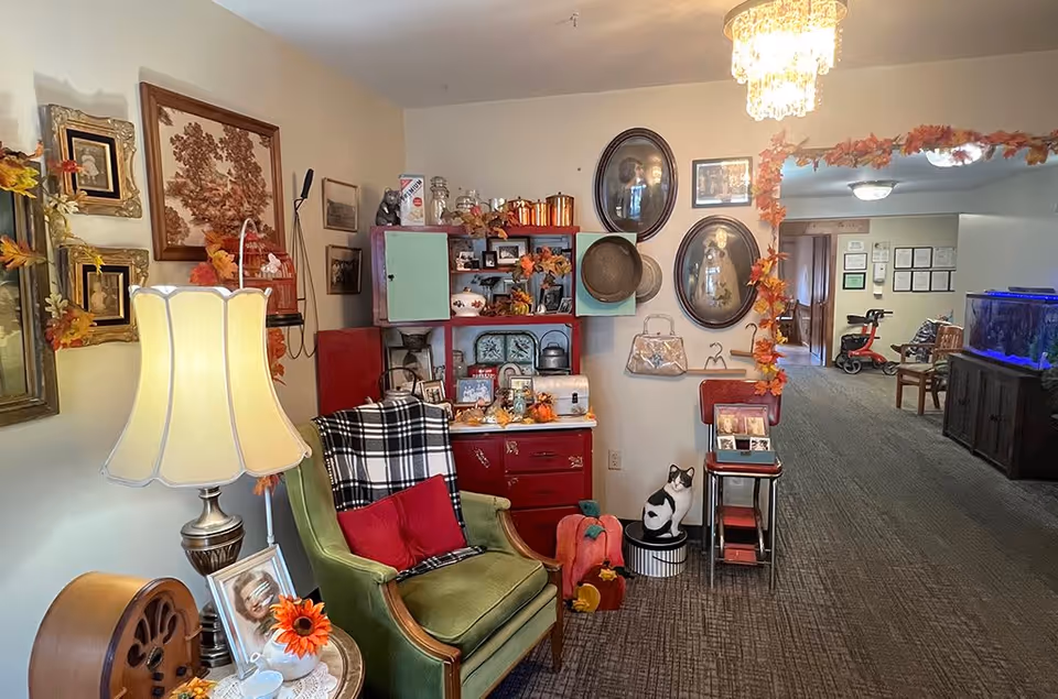A cozy interior corner of a senior living facility decorated with vintage furniture and autumn-themed decor. There is a green armchair with red pillows and a black and white plaid blanket, a side table with a lamp, framed pictures on the wall, and a red cabinet with various decorative items. The hallway extends to the right with additional seating and a large fish tank.