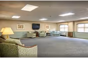 A spacious living room area with carpeted floor, several upholstered armchairs and sofas arranged around the room, a wall-mounted flat screen TV, two framed pictures on the wall, and large windows letting in natural light.