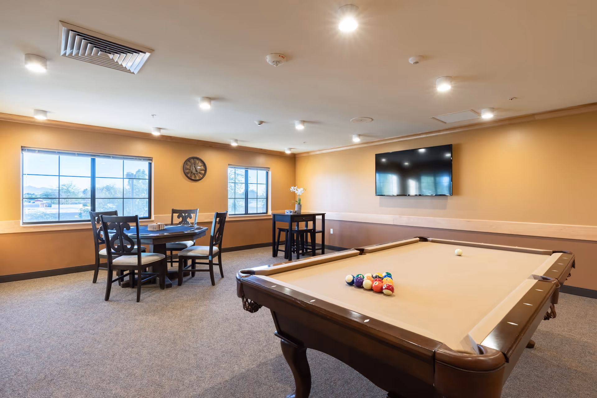 A spacious recreational room with a pool table in the foreground, a round table with four chairs near two large windows, a wall-mounted TV, and warm beige walls with ceiling lights.