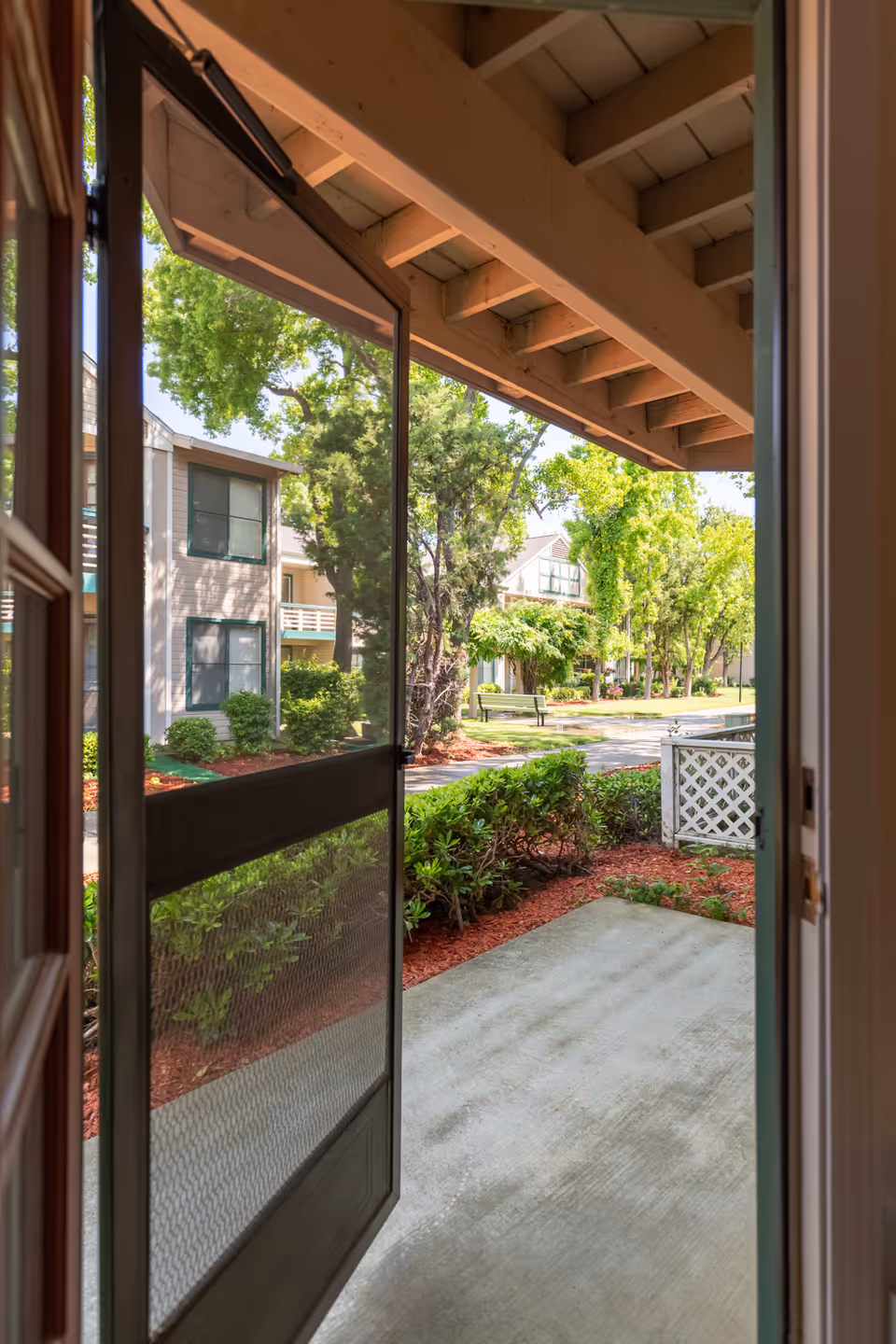 View through an open screen door looking out onto a small concrete patio area with landscaped bushes and red mulch. Beyond the patio, there are trees, shrubs, and a pathway with benches, as well as residential buildings in the background under a clear sky.