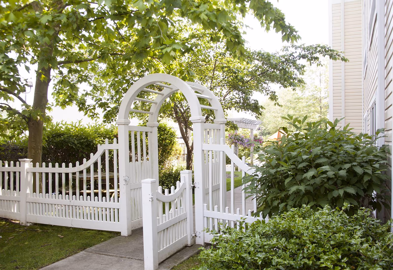 A white wooden garden gate with an arched trellis surrounded by green bushes and trees, next to a beige building exterior, leading to a garden path.