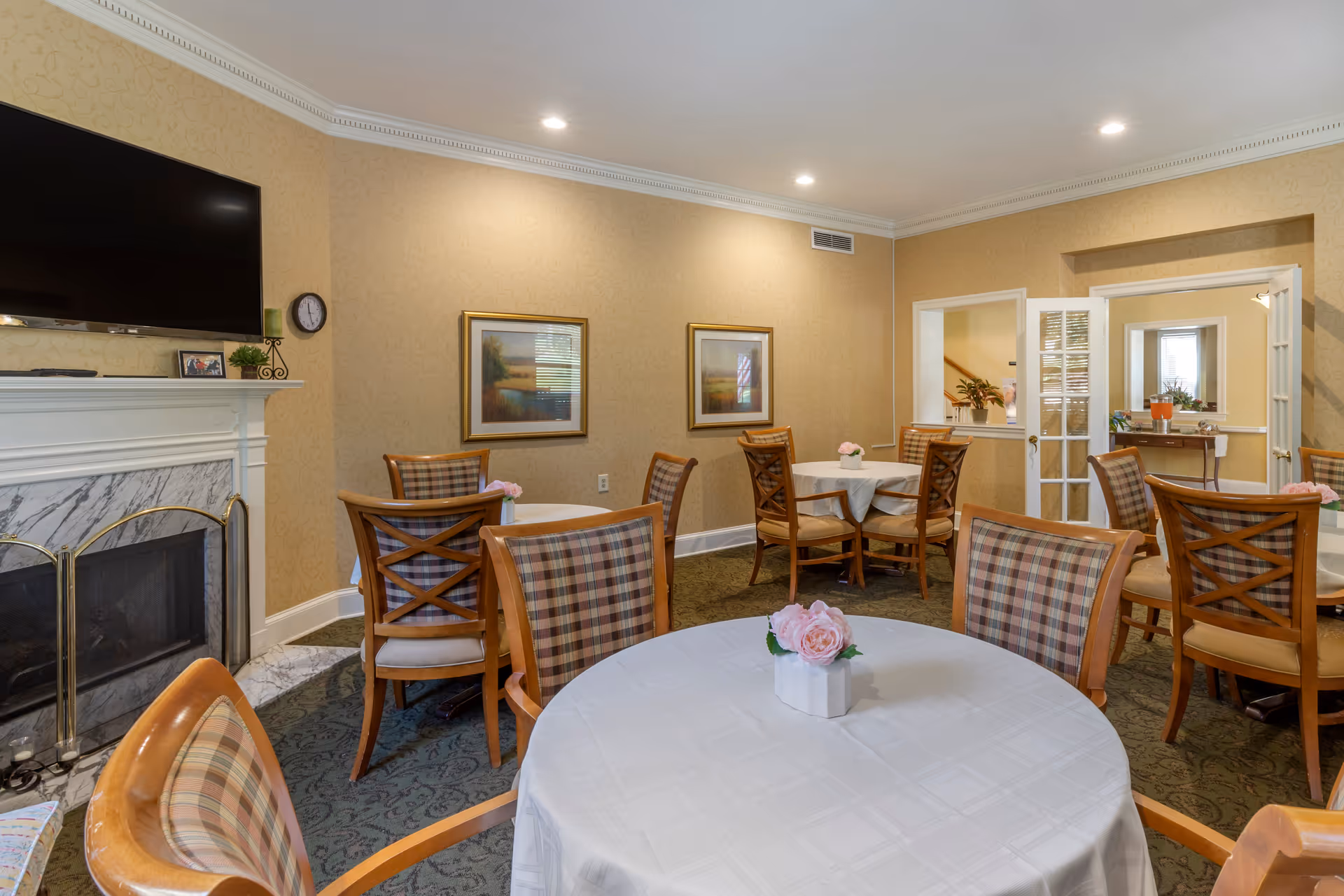 A cozy dining room with round tables covered in white tablecloths, each adorned with a small vase of pink flowers. The room features wooden chairs with plaid cushions, beige patterned wallpaper, a marble fireplace with a mounted flat-screen TV above it, and framed landscape paintings on the walls. There is a doorway with glass-paneled double doors leading to another room.