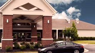 Front exterior view of Providence Assisted Living facility showing a covered entrance with brick pillars, a parked black car in front, and a partly cloudy sky.