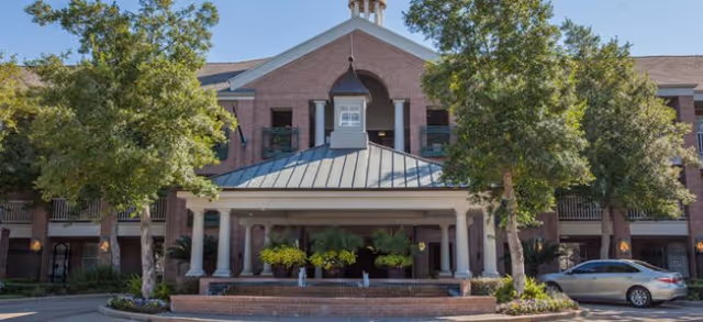 Front exterior view of a large brick building with a covered entrance supported by white columns, surrounded by trees and landscaping, with a car parked on the right side.