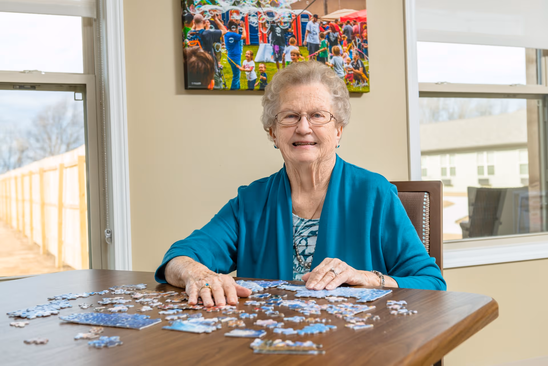 An elderly woman with glasses and a teal cardigan is sitting at a wooden table working on a jigsaw puzzle. Behind her is a beige wall with a colorful photo of people at an outdoor event. There are two windows on either side of her showing an outdoor view with a fence and buildings.