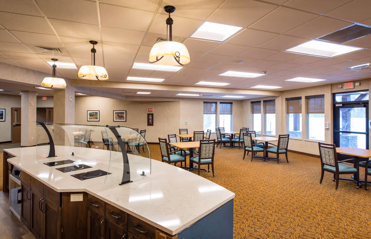 A spacious dining area in a senior living facility with multiple tables and chairs arranged neatly. The room features large windows with blinds, beige walls, and a patterned carpet. In the foreground, there is a serving counter with a glass sneeze guard and overhead pendant lights.