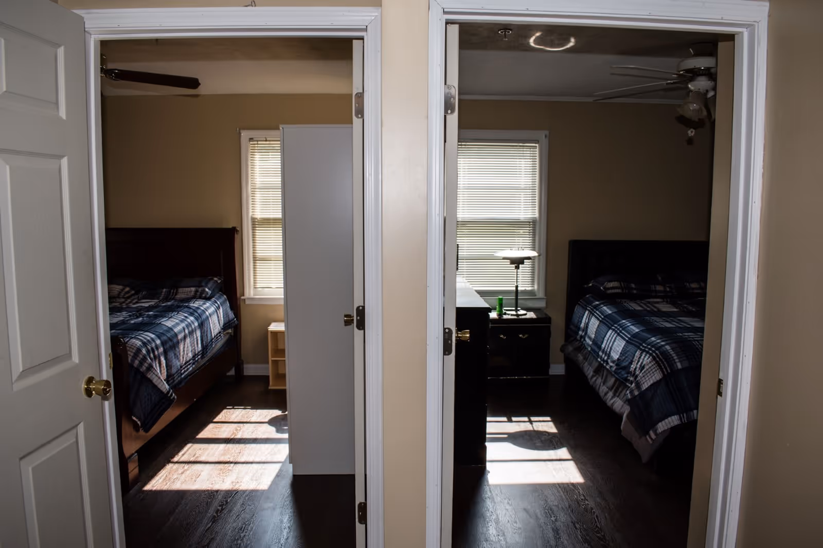 View of two adjacent bedrooms separated by a wall, each with a bed covered in blue plaid bedding, a window with blinds, and a nightstand. The left bedroom has a ceiling fan and a small wooden nightstand, while the right bedroom has a ceiling fan, a lamp, and a dark nightstand.