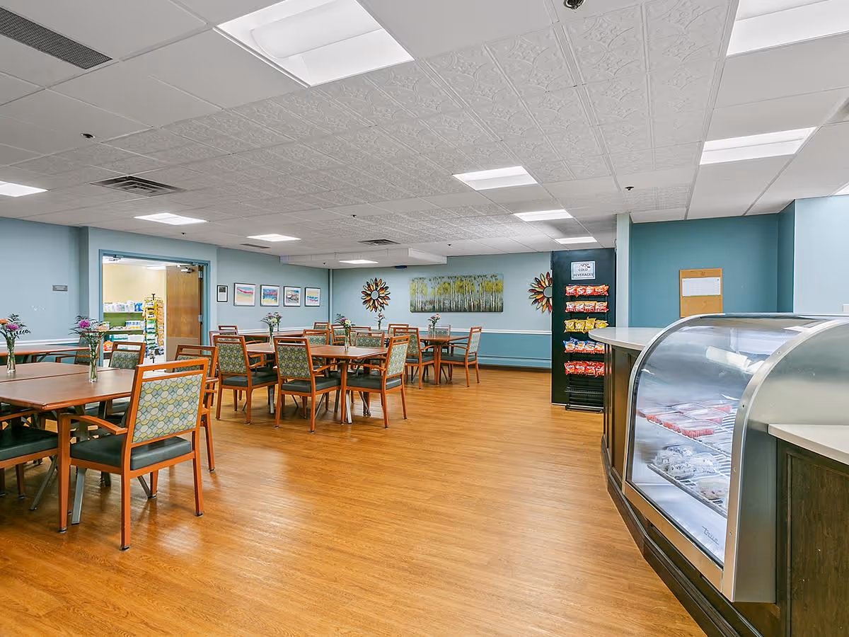 A spacious dining room with multiple wooden tables and chairs arranged neatly. Each table has a small vase with flowers. The walls are painted light blue and decorated with framed pictures and colorful wall art. On the right side, there is a refrigerated display case with food items and a rack stocked with bags of chips and cold beverages. The floor is wooden, and the ceiling has white tiles with recessed lighting.