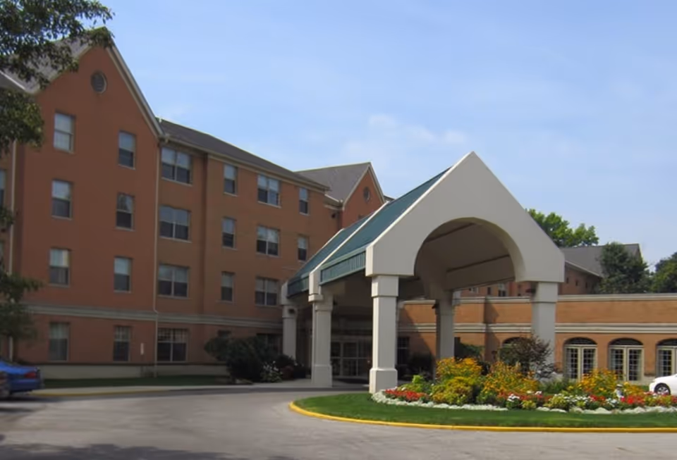 Exterior view of a multi-story brick building with a covered entrance and a landscaped roundabout with flowers and greenery in front.