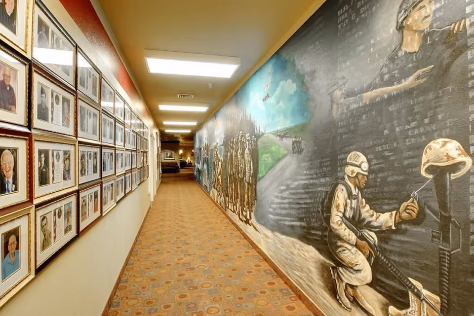 A long hallway with framed photographs on the left wall and a large mural depicting soldiers and military scenes on the right wall. The floor is carpeted with a patterned design, and the ceiling has rectangular fluorescent lights.