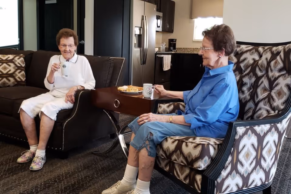 Two elderly women sitting in a cozy living room area, each holding a mug. One woman is seated on a dark brown sofa with patterned cushions, wearing a white outfit and colorful shoes. The other woman is seated on a patterned armchair, wearing a blue shirt and denim capris. A small wooden table between them holds a plate of pastries. In the background, a kitchen area with stainless steel appliances and granite countertops is visible.