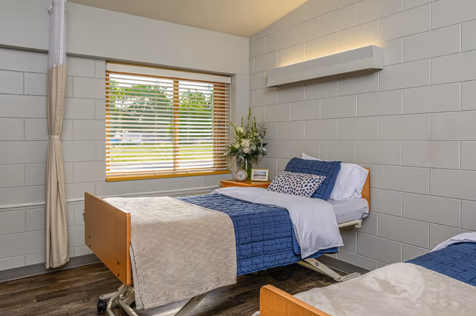 Two neatly made adjustable beds with blue bedding in a simple room with a window, nightstand and floral arrangement.