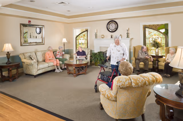 A cozy senior living facility common area with several elderly residents sitting and chatting. The room features comfortable upholstered chairs and sofas, a coffee table, side tables with lamps, stained glass windows, and a white fireplace with decorative items on the mantel. One elderly woman is standing with a walker, engaging with another seated resident.