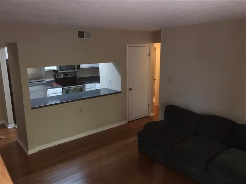 Interior view of a living area with a dark-colored couch on the right side and a pass-through window opening into a kitchen with white cabinets and stainless steel appliances. The floor is wooden, and there is a closed white door next to the kitchen opening.