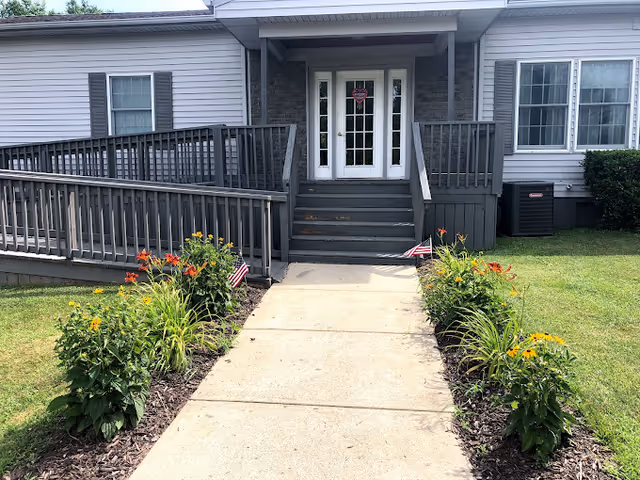 Entrance to a building with a concrete walkway leading up to stairs and a wheelchair ramp on the left side. The building has gray siding, white framed windows, and a white door with glass panels. There are flower beds with orange and yellow flowers along both sides of the walkway, and small American flags are placed in the flower beds.