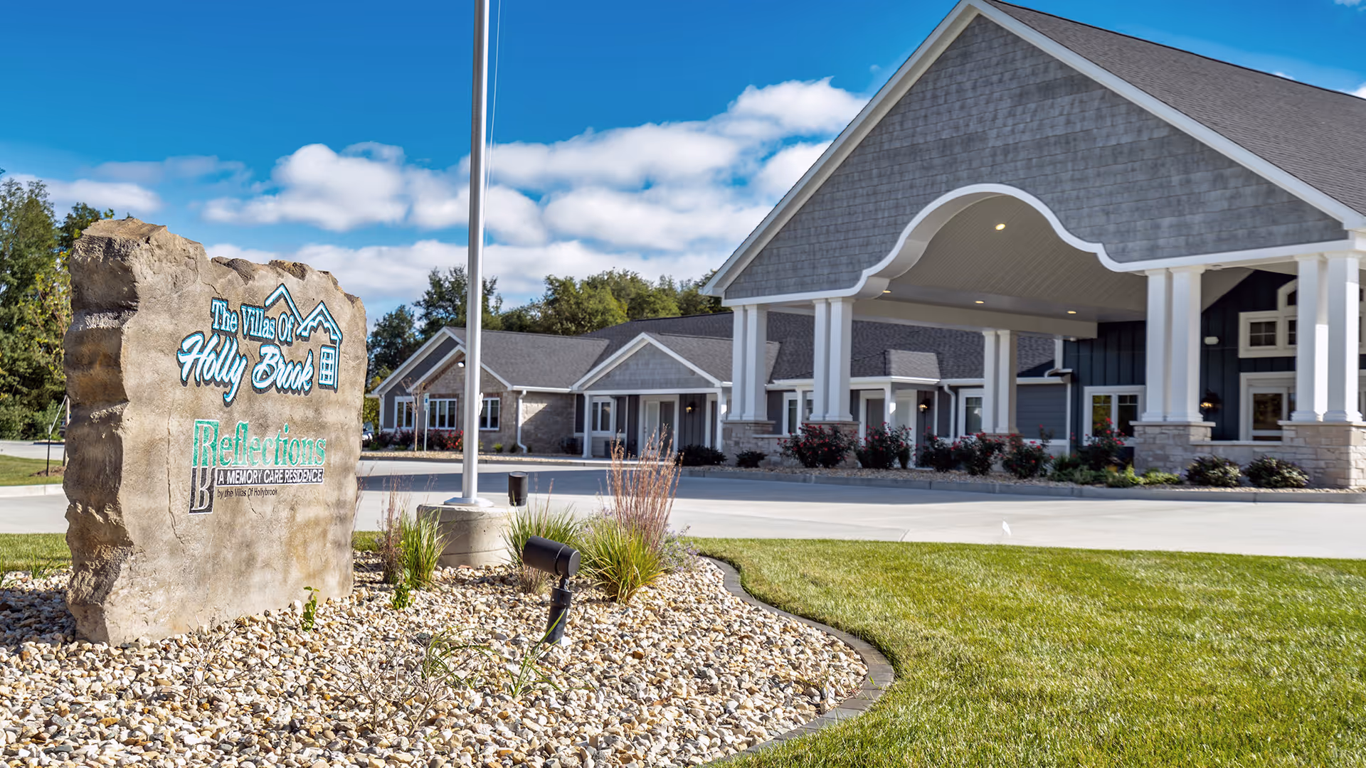 Front exterior view of Villas of Holly Brook Assisted Living & Memory Care facility with a large stone sign in the foreground and a covered entrance to the building under a blue sky with some clouds.