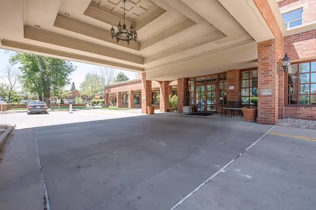 Covered entrance area of a senior living facility with brick pillars and walls, a chandelier hanging from the ceiling, potted plants, and glass doors leading inside. A car is parked outside near trees and greenery.